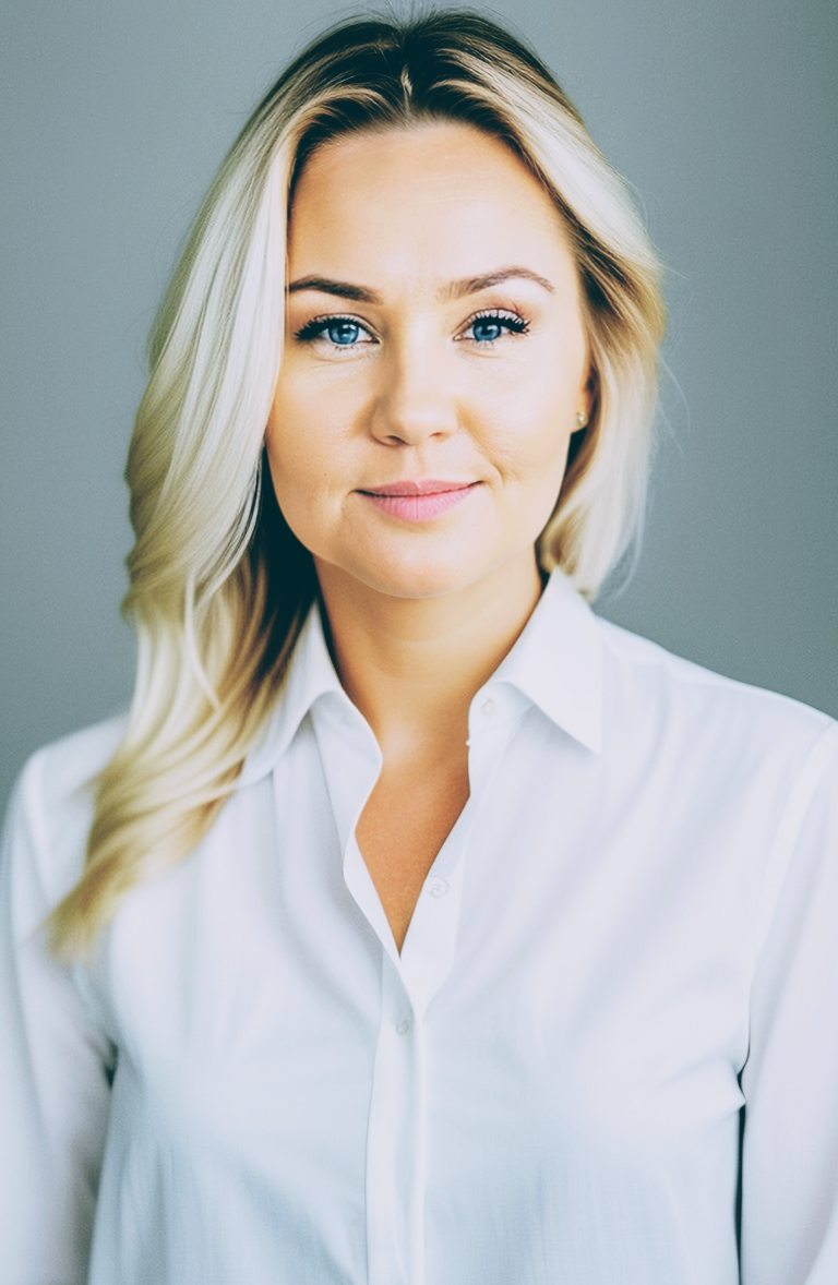 Portret studyjny uśmiechniętej młodej kobiety o blond włosach i niebieskich oczach, ubranej w białą koszulę z kołnierzykiem, patrzącej prosto w obiektyw. Tło jest gładkie i szare.