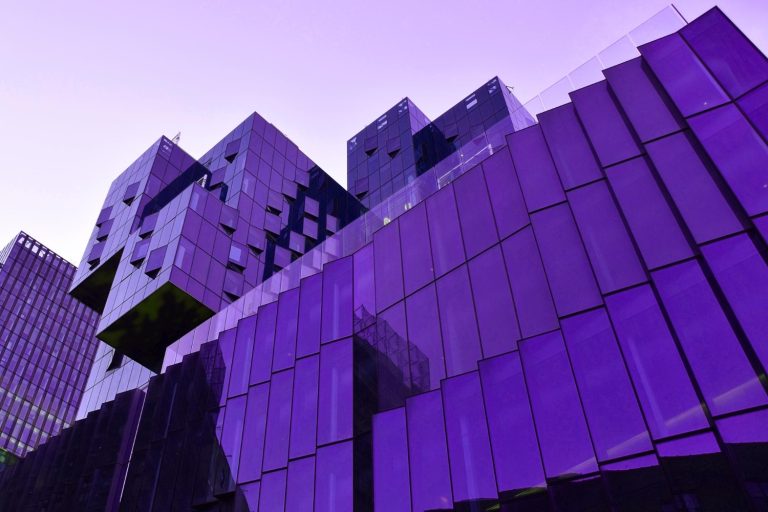 Architektoniczny widok z dołu na nowoczesny budynek biurowy, z dominującymi fioletowymi i ciemnoniebieskimi szklanymi fasadami. Elewacja składa się z nieregularnych, geometrycznych paneli, nadających budynkowi futurystyczny wygląd.